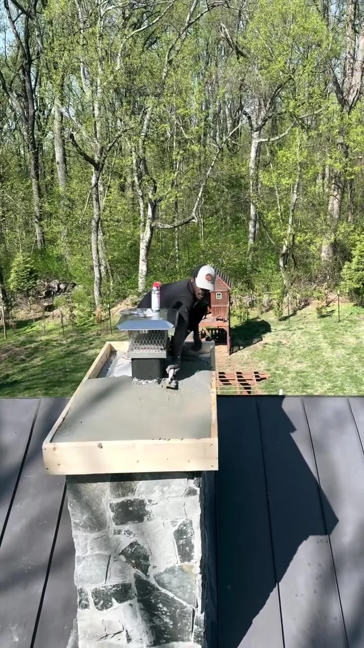 Masonry technician working on a stone chimney top above a dark metal roof